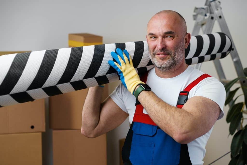 Man mover in uniform carrying big rolled carpet for relocation